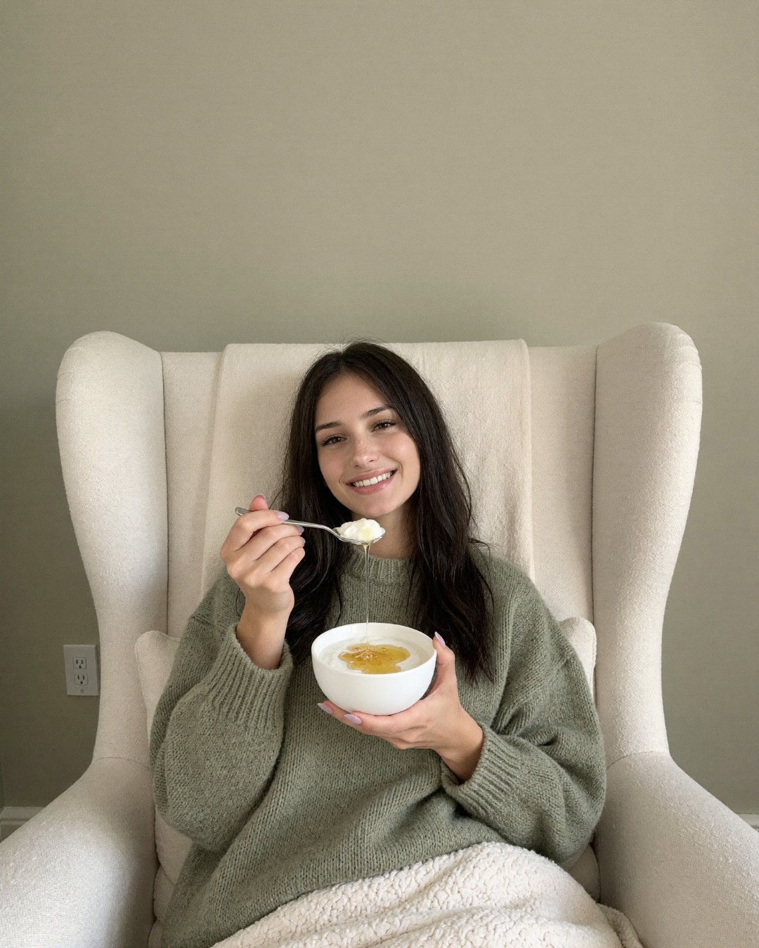 A woman curled up in a cream armchair under a blanket, holding a small bowl of yogurt with honey